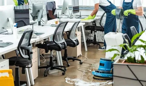 addetto alle pulizie che spolvera le scrivanie di un ufficio mentre una macchina per la pulizia del pavimento viene passata da un altro addetto addetto alle pulizie che spolvera le scrivanie di un ufficio mentre una macchina per la pulizia del pavimento viene passata da un altro addetto